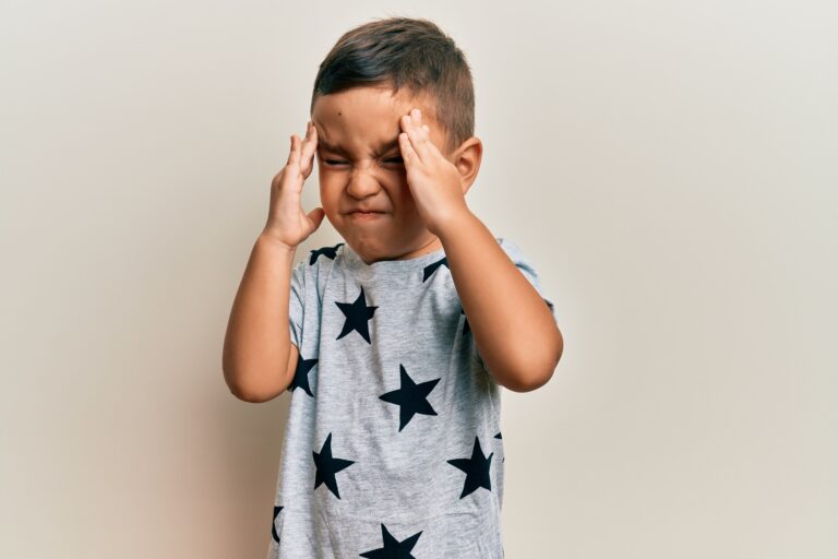 Adorable latin toddler wearing casual clothes with hand on head, headache because stress. suffering migraine. Frustrated young boy with closed eyes and hands on head, expressing distress—illustrating emotional overwhelm and meltdowns in autistic children, relevant to parenting strategies and autism support.