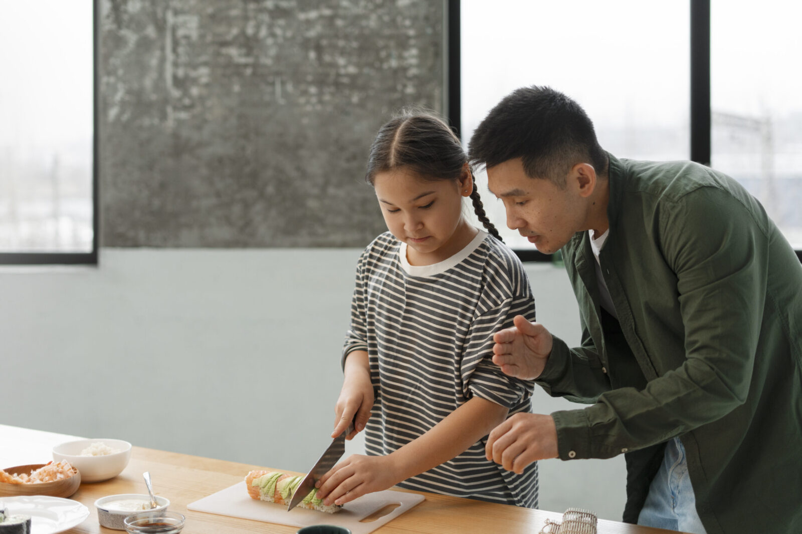 A father guides his daughter as she carefully cuts sushi rolls on a kitchen counter, illustrating hands-on learning and skill-building. A concept relevant to vocational training for teens with autism.