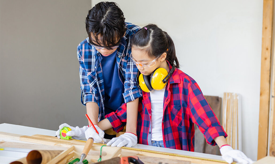 A mentor wearing glasses and a plaid shirt guide a younger girl wearing safety goggles