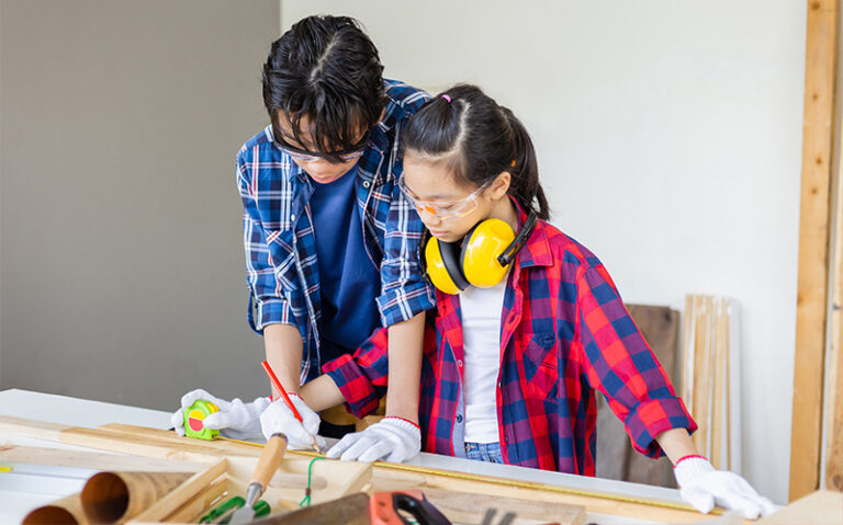 A mentor wearing glasses and a plaid shirt guide a younger girl wearing safety goggles A mentor wearing glasses and a plaid shirt guide a younger girl wearing safety goggles