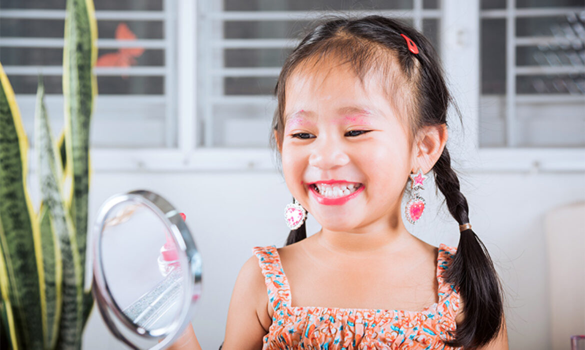 A young girl with braided hair and colorful earrings smiling at her reflection