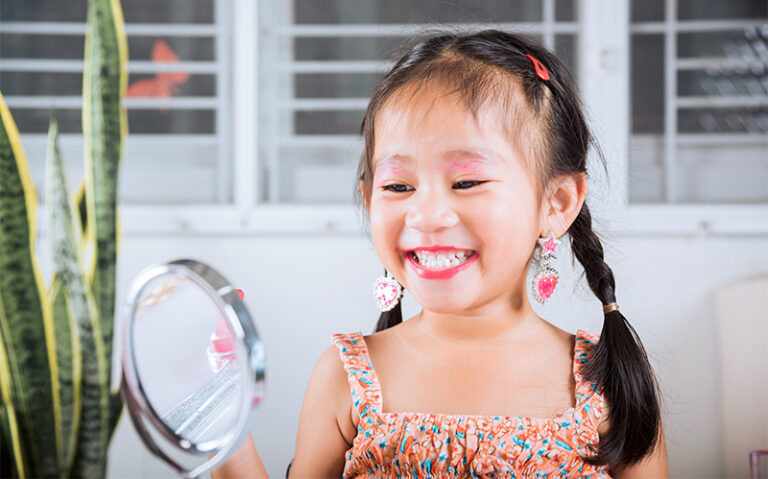 A young girl with braided hair and colorful earrings smiling at her reflection